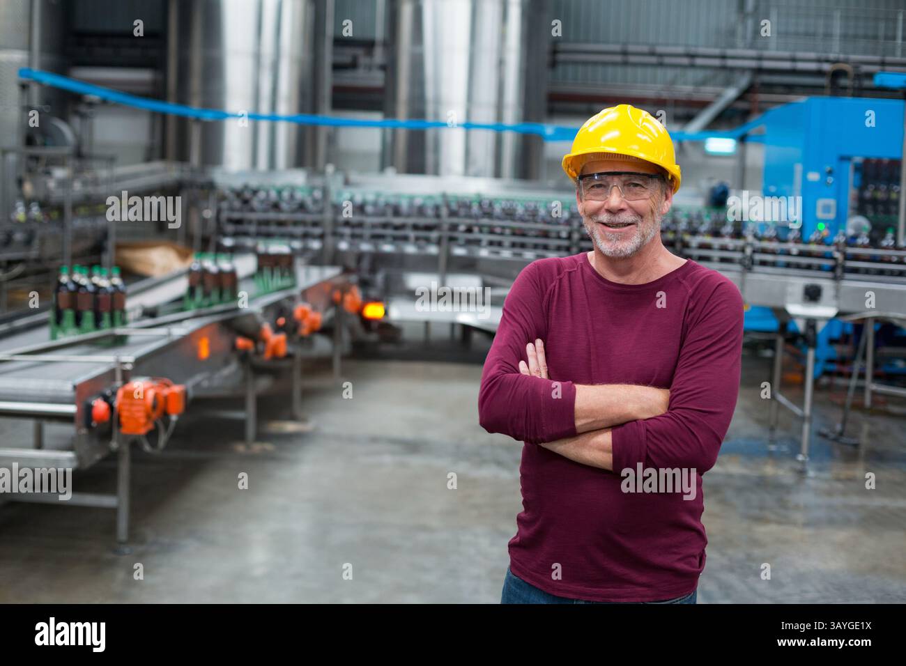 Lavoratore anziano che indossa un elmetto giallo che ispeziona il trasportatore di bottiglie verdi in fabbrica, spazio di copia Foto Stock