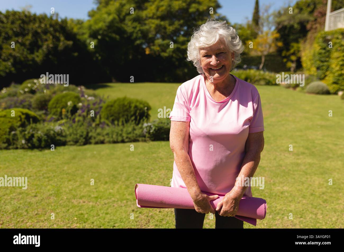 Donna anziana in piedi sul prato nel cortile con siepi rifinite, tenendo in mano un tappetino da yoga rosa arrotolato Foto Stock