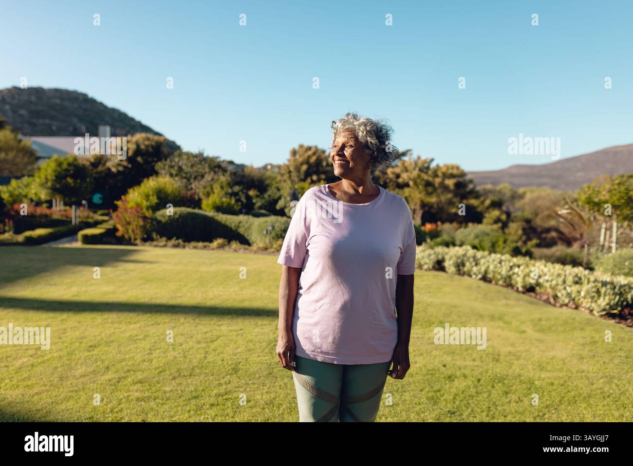 Donna anziana in piedi e che guarda a sinistra nel giardino residenziale, godendo del sole e della vegetazione lussureggiante Foto Stock