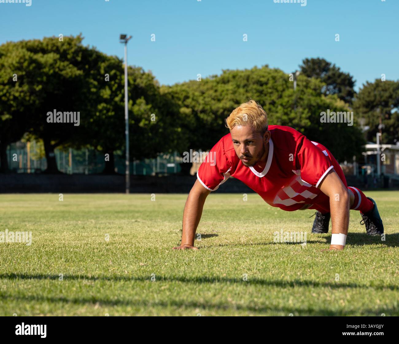 Uomo che esegue push-up su un campo sportivo in erba, indossa braccialetti bianchi in maglia rossa e tacchetti neri Foto Stock Uomo che esegue push-up su un campo sportivo in erba, indossa braccialetti bianchi in maglia rossa e tacchetti neri Foto Stock