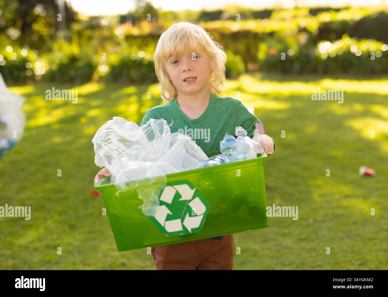 Bambino che tiene in mano un cestino per il riciclaggio verde in un cortile illuminato dal sole, smistamento di bottiglie di plastica e sacchetti trasparenti Foto Stock