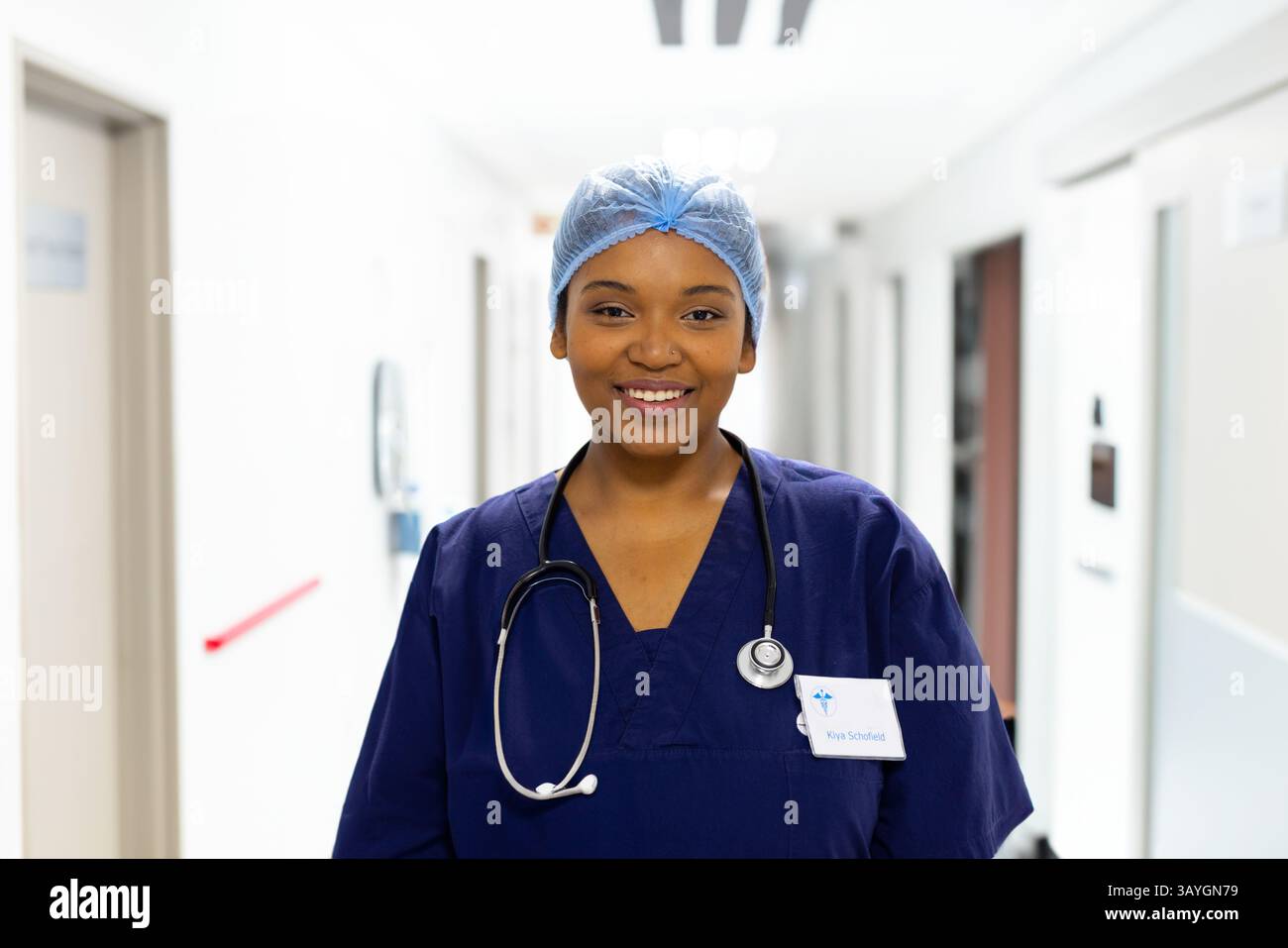 Infermiera sorridente e in piedi in corridoio ospedale, indossando cappello chirurgico, stetoscopio, badge ID Foto Stock