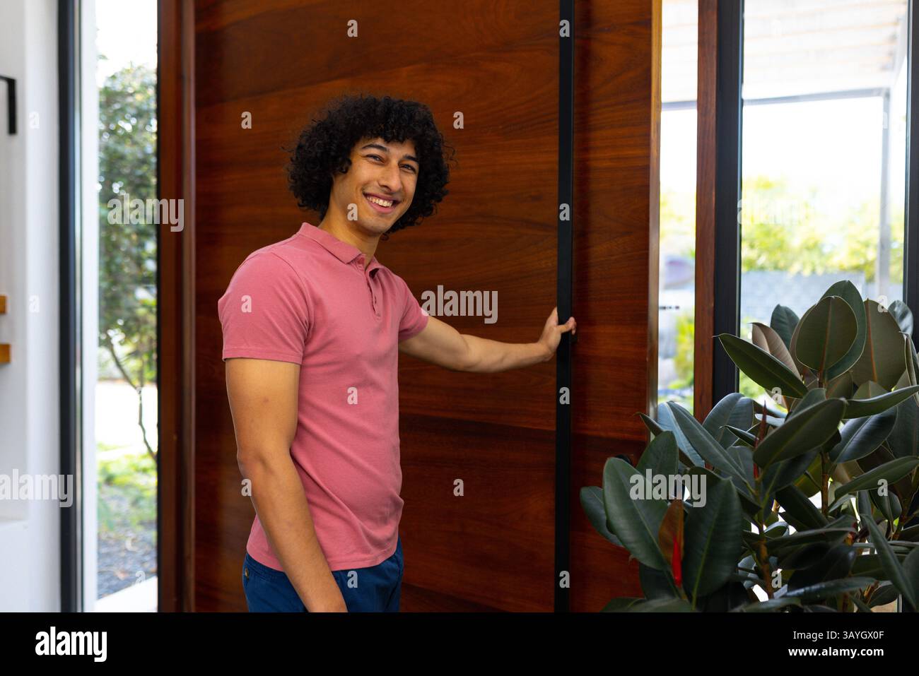 Uomo eterogeneo che apre la porta girevole nell'ingresso della casa con pianta in gomma e finestra in vetro che mostra il giardino Foto Stock
