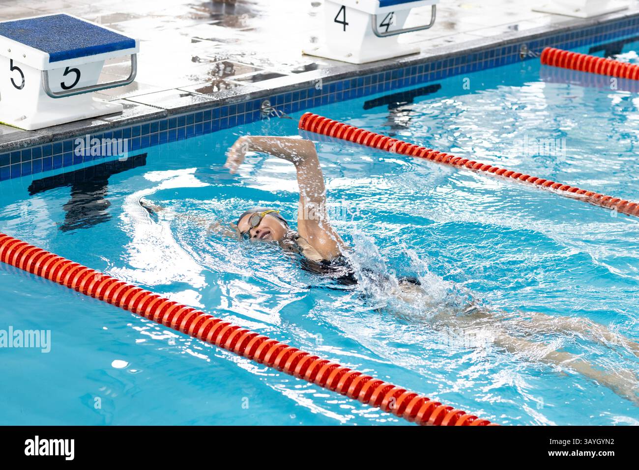 Nuotatrice adolescente che esegue un colpo di schiena in piscina, indossando un cappello giallo e occhiali di protezione Foto Stock