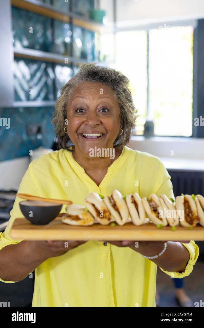 Donna anziana che presenta un tavolo da servizio in legno con panini cotti al vapore e salsa per immersione nella cucina casalinga Foto Stock