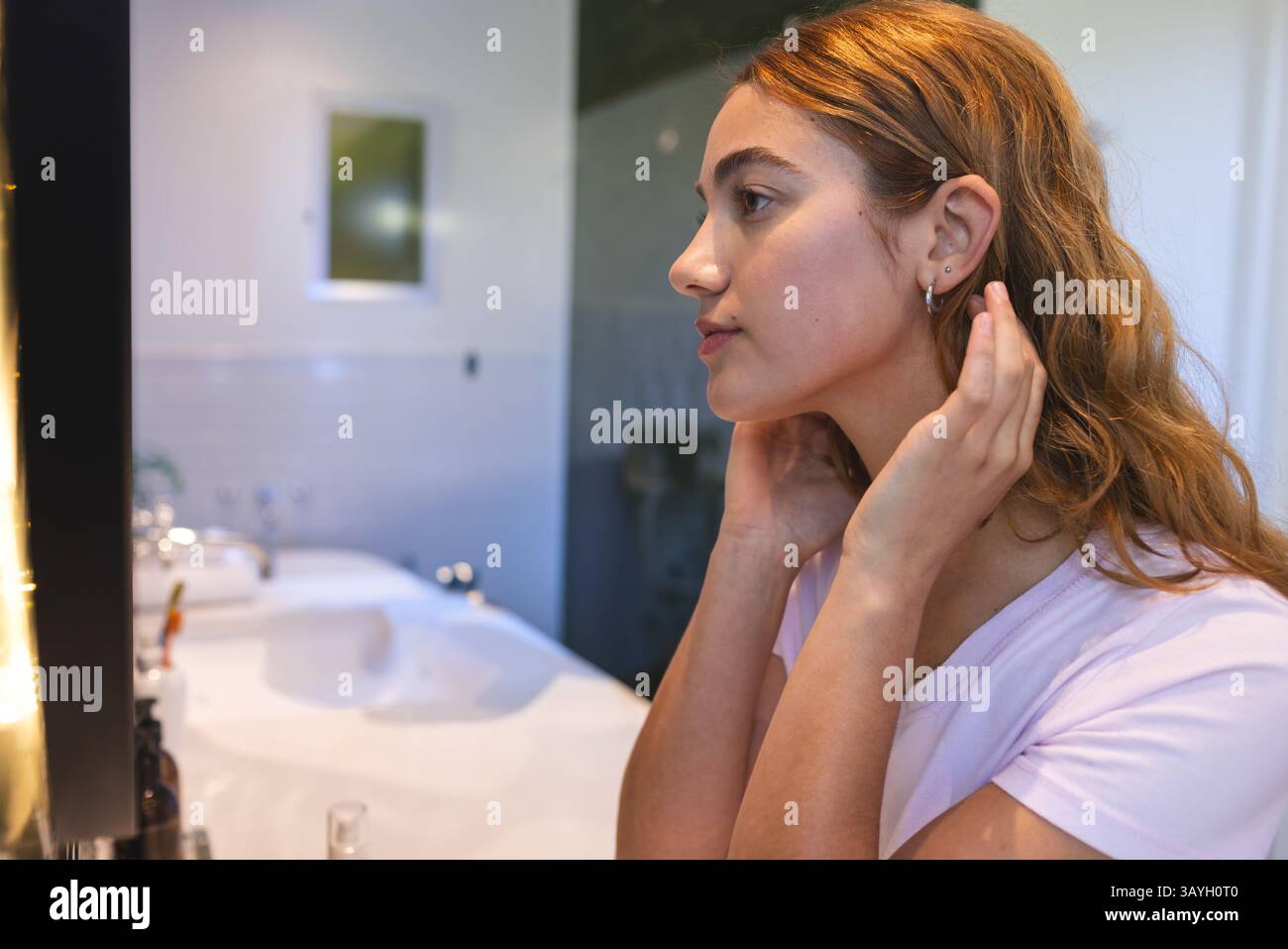 Donna che regola orecchini e capelli in piega nel bagno di casa, con specchio per lavello e bottiglie per articoli da toeletta Foto Stock