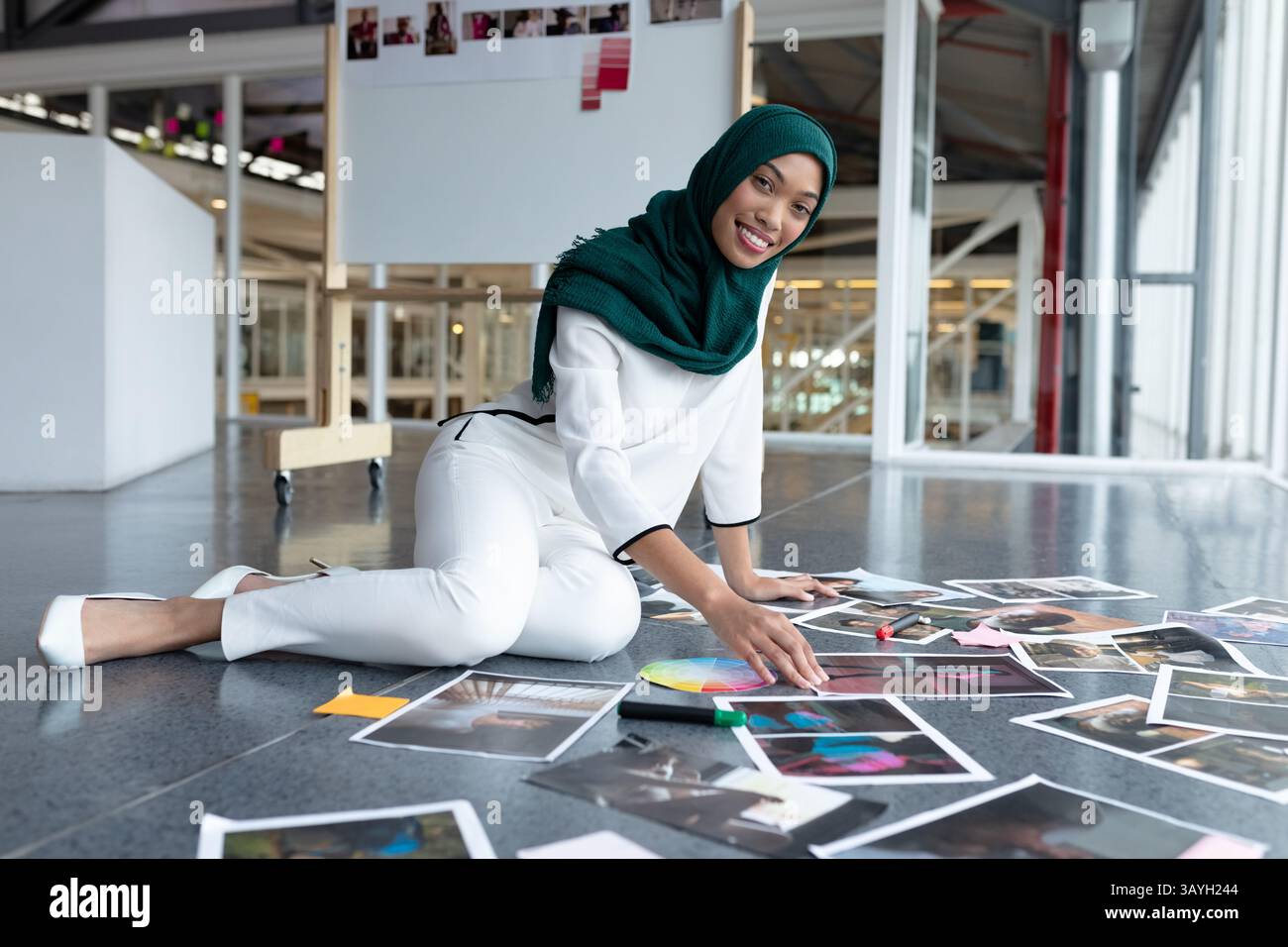 Donna che indossa un hijab verde inginocchiato e organizza fotografie e campioni sul moderno piano di studio Foto Stock