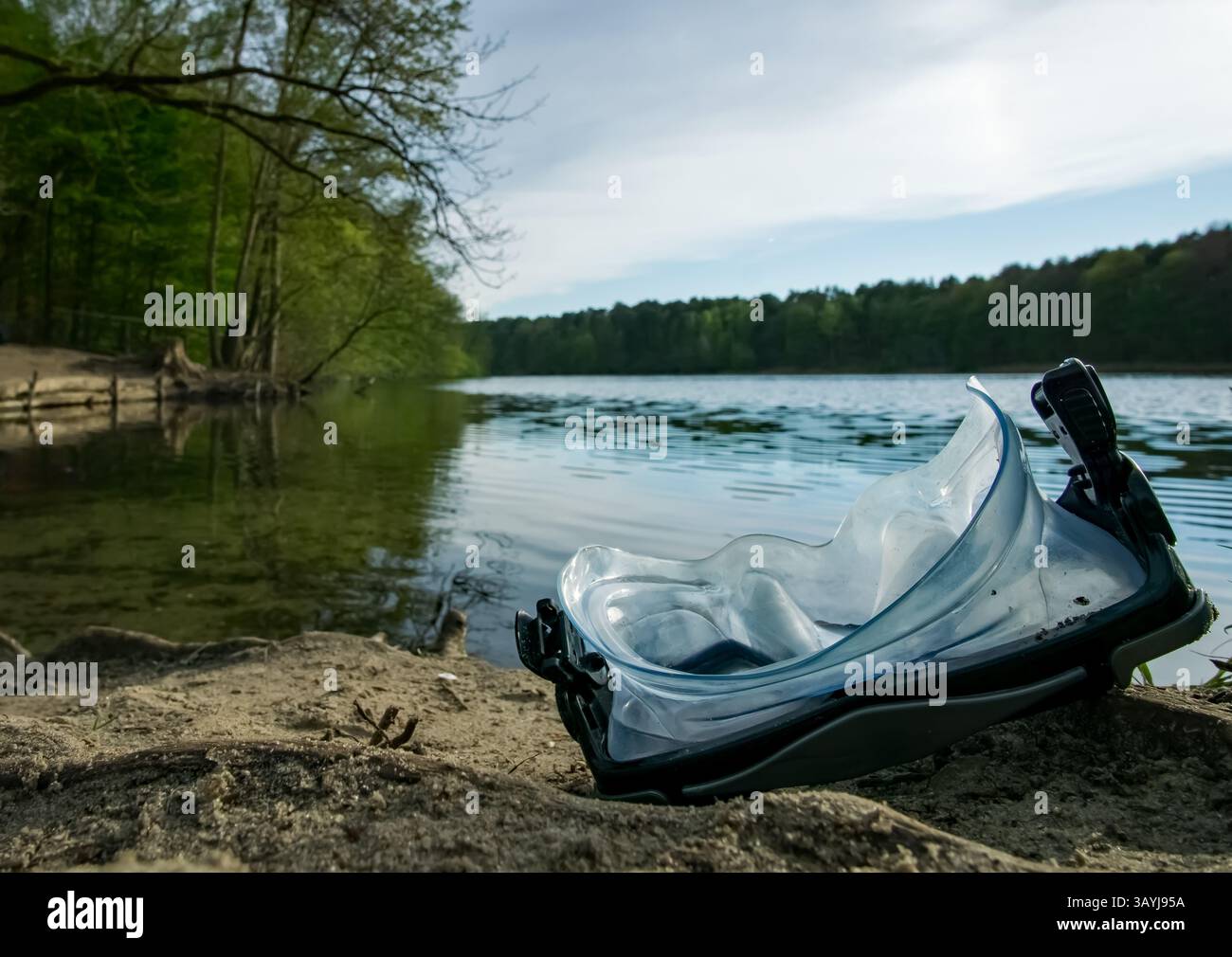 Una maschera subacquea poggia sulla riva sabbiosa del lago Krumme Lanke a Berlino, con acque calme e foresta sullo sfondo. Foto Stock