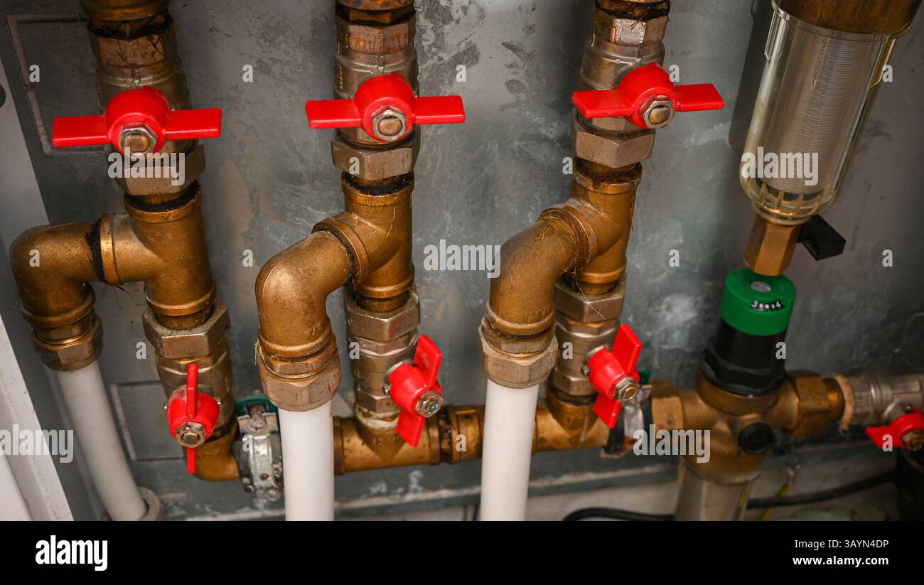 Primo piano di tubi idraulici in rame con valvole rosse e un filtro, collegati a tubi bianchi, all'interno di un locale caldaia. Foto Stock
