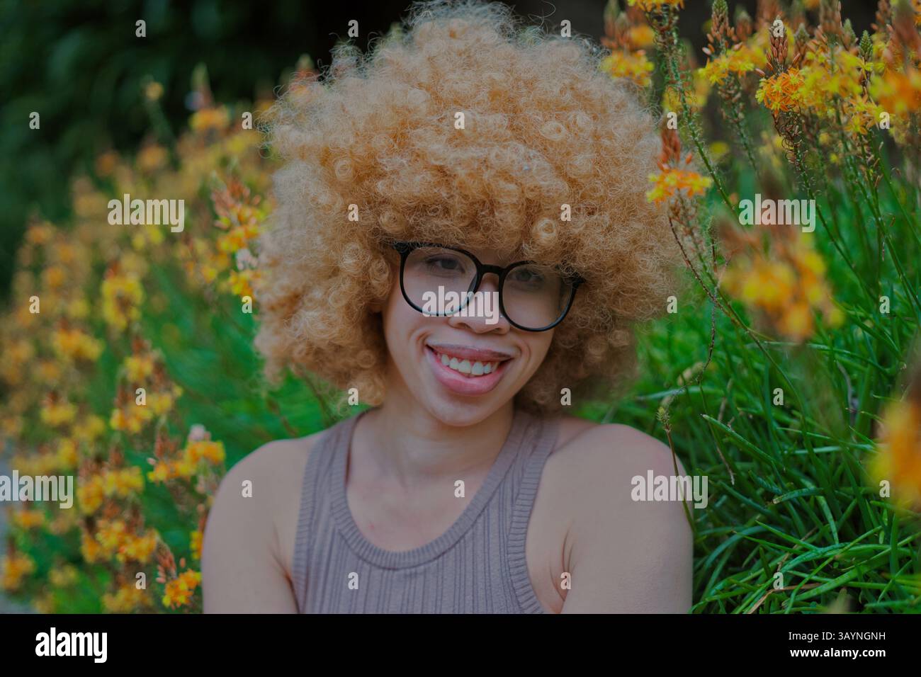 Giovane donna dai capelli afro biondi sorridente in un giardino con fiori gialli Foto Stock