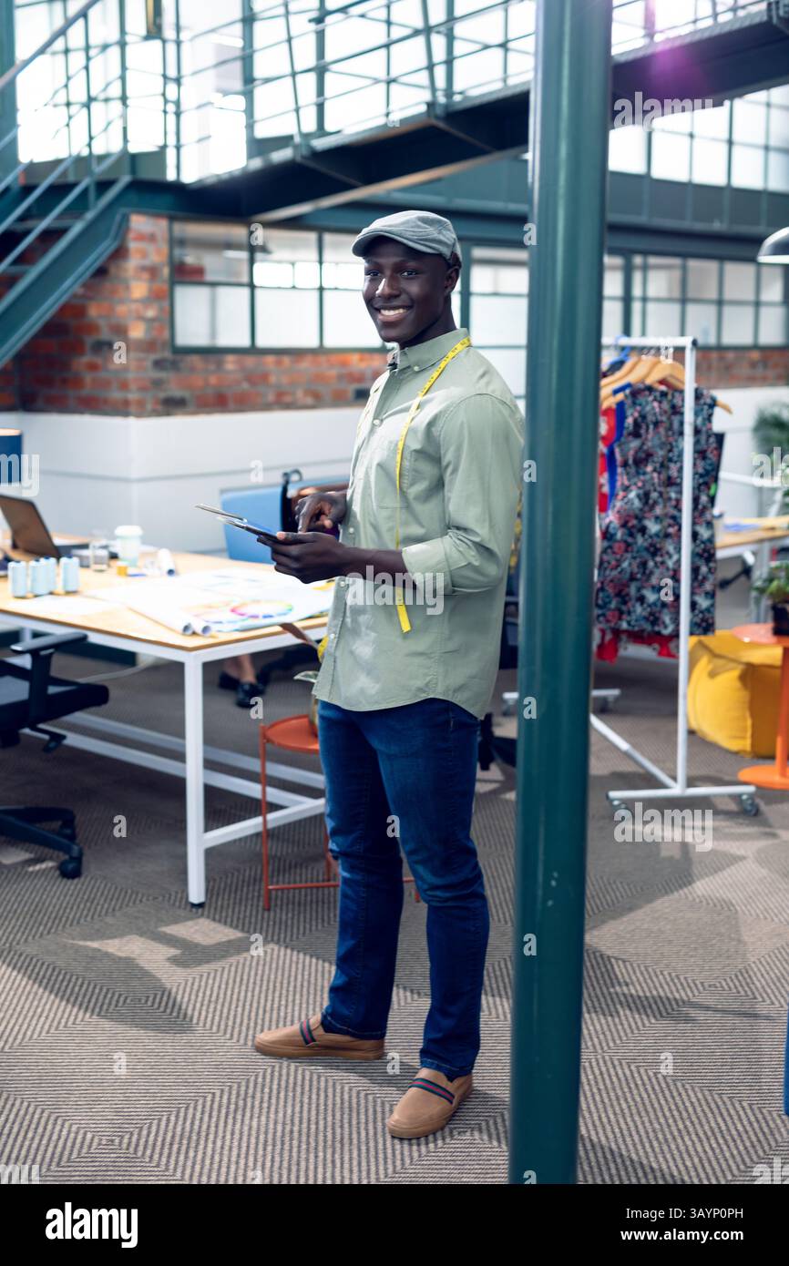 Uomo afroamericano che rivede i progetti su tablet digitale con metro a nastro nello spazio di lavoro della moda Foto Stock