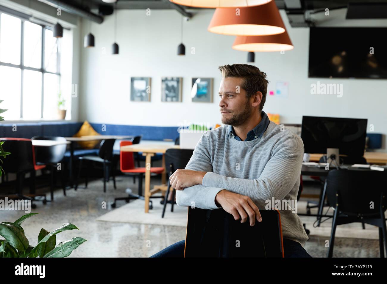 Uomo seduto su una sedia da ufficio all'indietro nello spazio di coworking, con luci sospese e spazio fotocopie Foto Stock