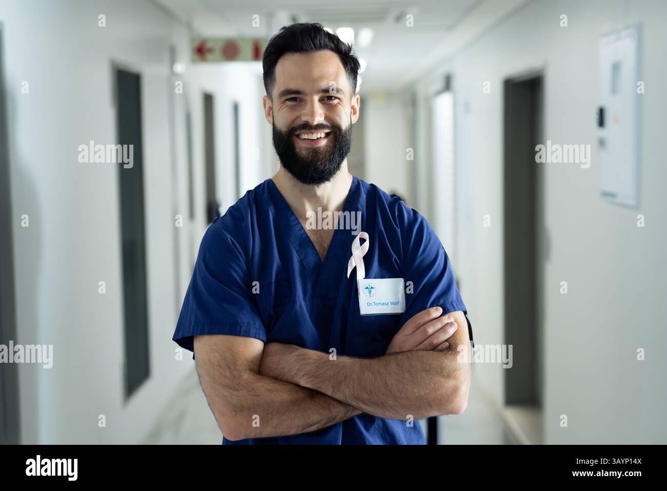 Giovane medico in posa e sorridente in corridoio ospedale con scrub blu con nastro e badge nome Foto Stock