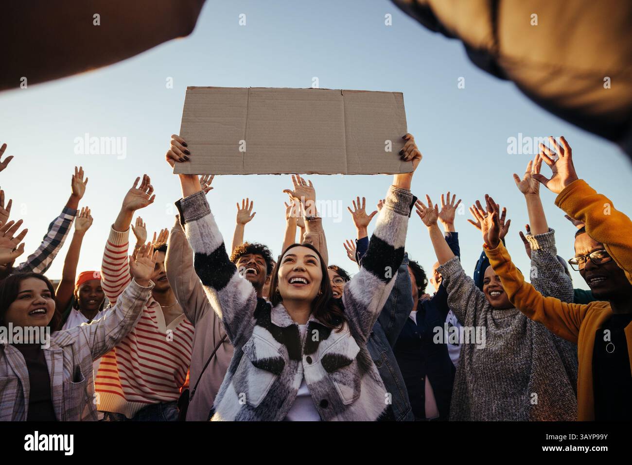 Un gruppo eterogeneo di persone si riunisce all'aperto per sostenere una causa, alzando le mani con entusiasmo mentre una giovane donna centrata tiene in alto un sig vuoto Foto Stock