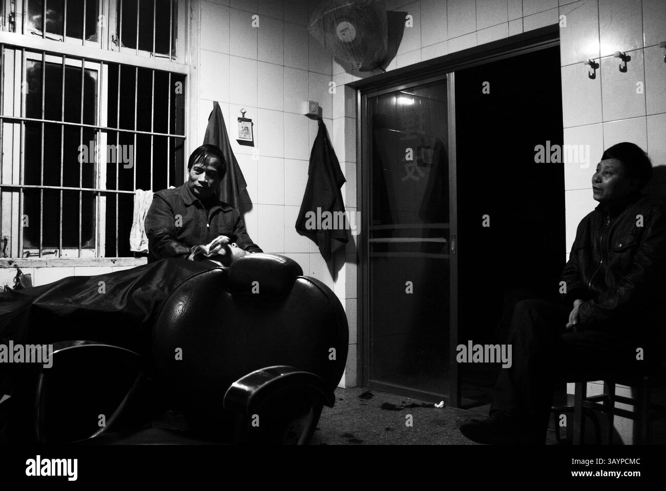 Il barbiere scambia in un ambiente intimo durante le ore serali: Barbiere barba del cliente nel barbiere cinese degli anni '2000 Foto Stock