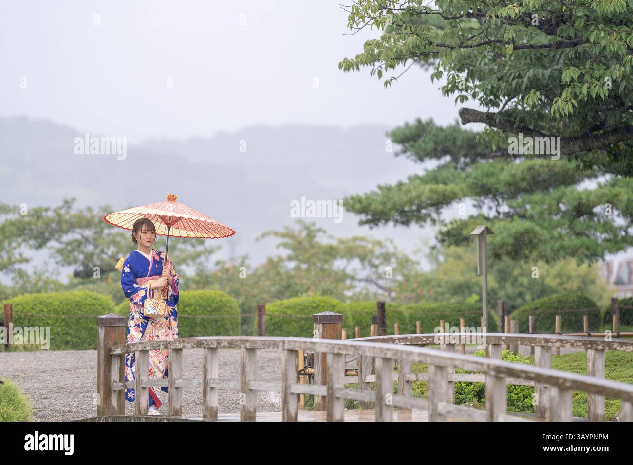 Una città locale in Giappone in un giorno d'estate piovoso. Donna giapponese che indossa un kimono adolescente blu con un ombrello giapponese che cammina lungo un giardino tradizionale Foto Stock