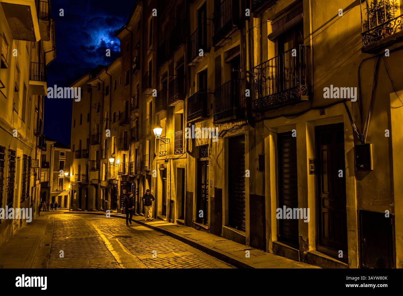 Una strada a Cuenca giace silenziosa al chiaro di luna mentre edifici e pochi pedoni costeggiano la Calle Santa Catalina nella notte del Venerdì Santo. Castilla-La Mancha, Spagna. Il vicolo lastricato del centro storico si trova tra due tappe della processione del Venerdì Santo Camino del Calvario. Durante la Semana Santa a Cuenca, 33 Hermandades partecipano a undici processioni attraverso la città durante la Settimana Santa. Foto Stock
