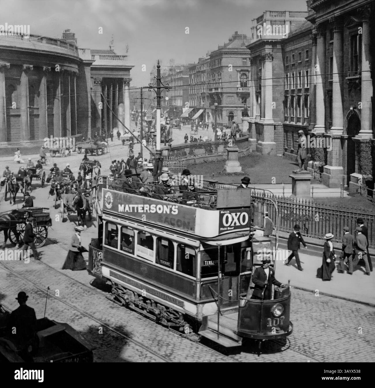 Centro di Dublino, Irlanda. Con persone, veicoli trainati da cavalli e tram elettrici di nuova introduzione. La sinistra è la banca d'Irlanda - una volta sede del Parlamento irlandese. Sulla destra c'è Regent House, che funge da ingresso centrale al campus del Trinity College di Dublino, Irlanda. Foto Stock