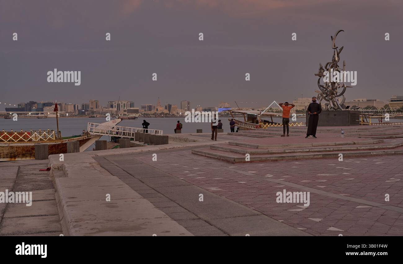 Skyline di Doha in Qatar dalla passeggiata in corniche foto al tramonto che mostra la scultura calligrafica e i dhows nel golfo arabo con bandiera del Qatar Foto Stock