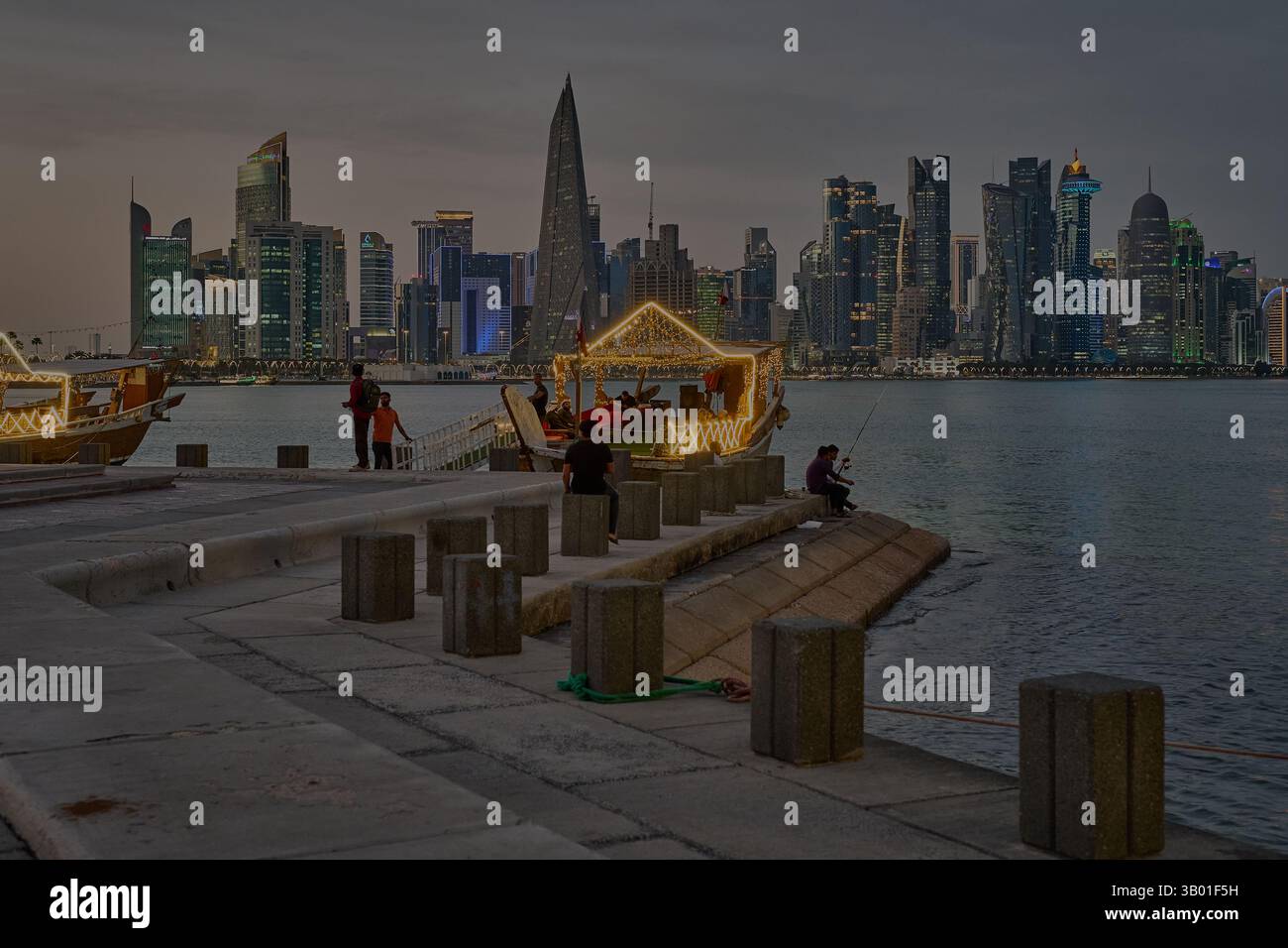 Skyline di Doha in Qatar dal tramonto sul lungomare di Corniche che mostra i dhows nel golfo arabo con la bandiera del Qatar Foto Stock