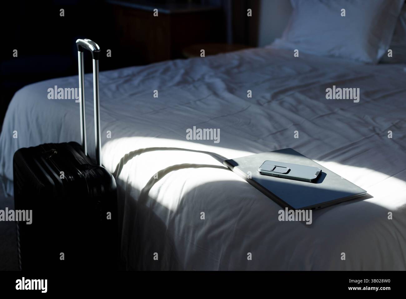 Valigia nera in piedi in camera da letto, che getta l'ombra della maniglia sul letto con il computer portatile e lo smartphone Foto Stock