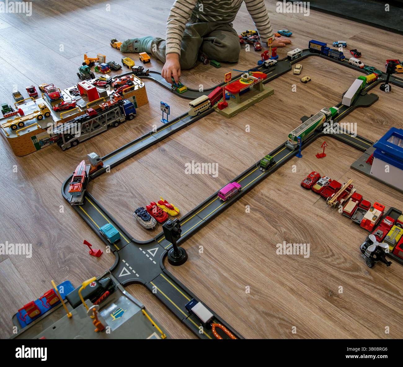 Bambino che gioca su un pavimento di legno con un'intricata installazione di macchinine giocattolo, strade ed edifici disposti in una configurazione simile a quella di una città Foto Stock