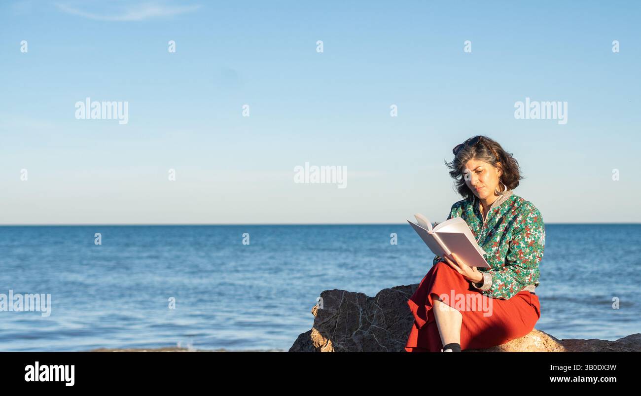Donna matura che legge un libro seduto su una roccia in riva al mare Foto Stock
