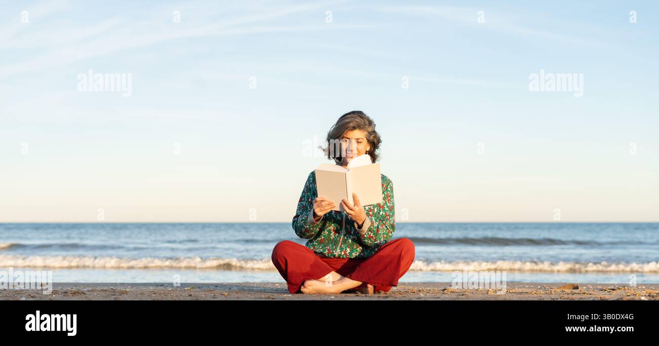 Donna matura che legge un libro mentre siede sulla spiaggia godendosi il tramonto Foto Stock