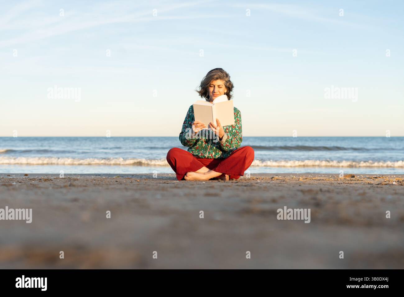 Donna anziana che si gode il suo tempo libero, leggendo un libro accattivante mentre si siede sulla spiaggia al tramonto Foto Stock