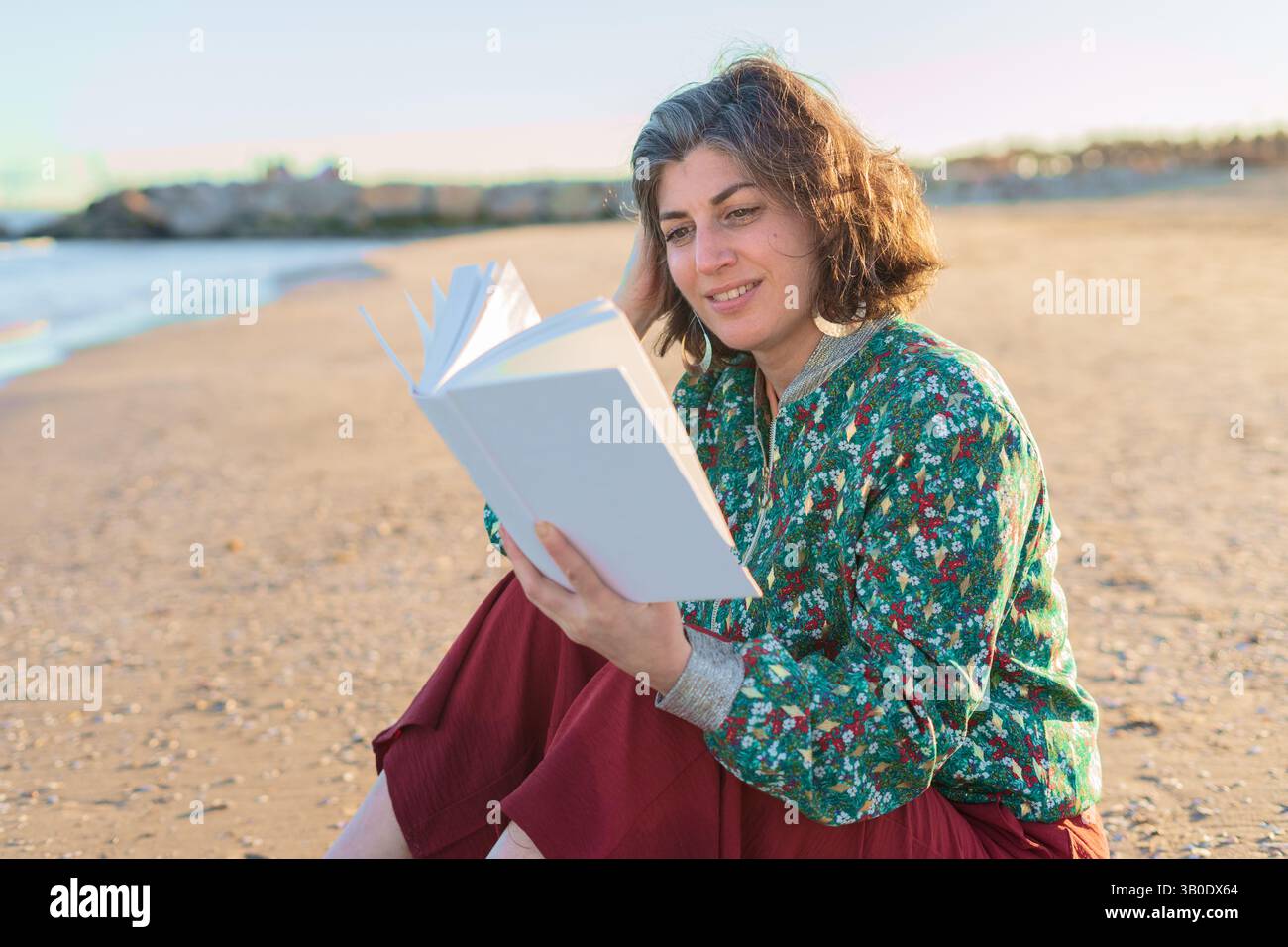Donna matura e felice seduta sulla spiaggia di sabbia, godendo un buon libro mentre si immerge nel caldo tramonto. Indossare abiti casual e sorridere, irradiare Foto Stock