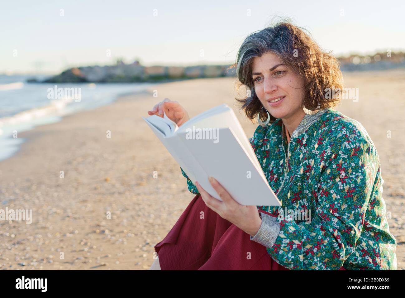 Donna matura sorridente con capelli grigi seduta sulla spiaggia, tenendo in mano un libro e godendosi la lettura durante un bellissimo tramonto, indossando un motivo verde Foto Stock