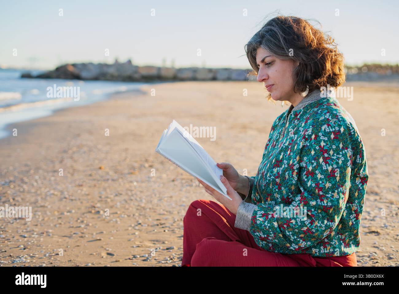 Donna di mezza età che si rilassa sulla spiaggia al tramonto, immersa in un libro mentre si siede sulla sabbia calda Foto Stock