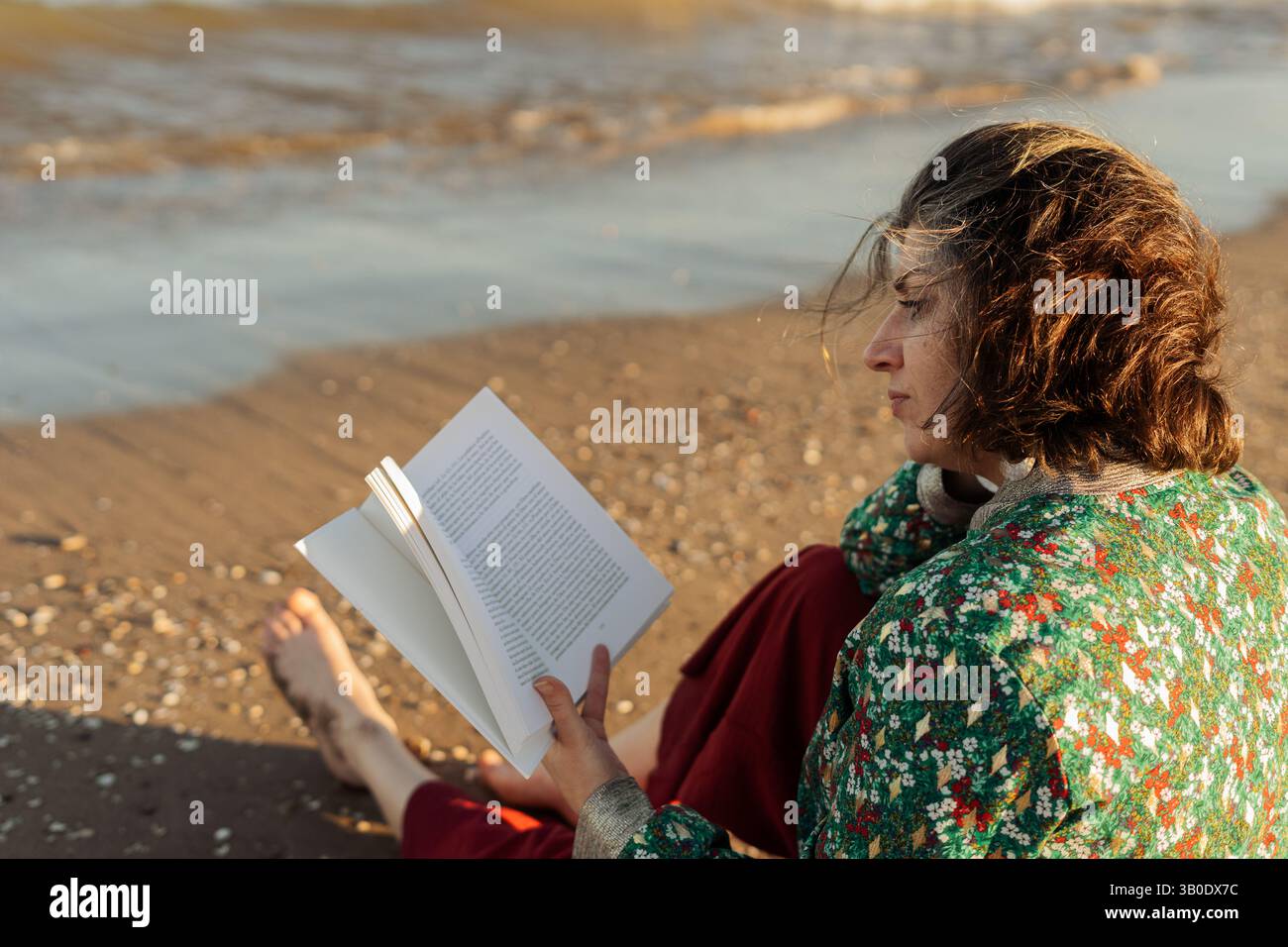 Giovane donna seduta a piedi nudi su una calda sabbia di spiaggia, immergendosi in un libro accattivante durante un momento tranquillo in riva al mare durante l'ora d'oro, abbracciando Foto Stock