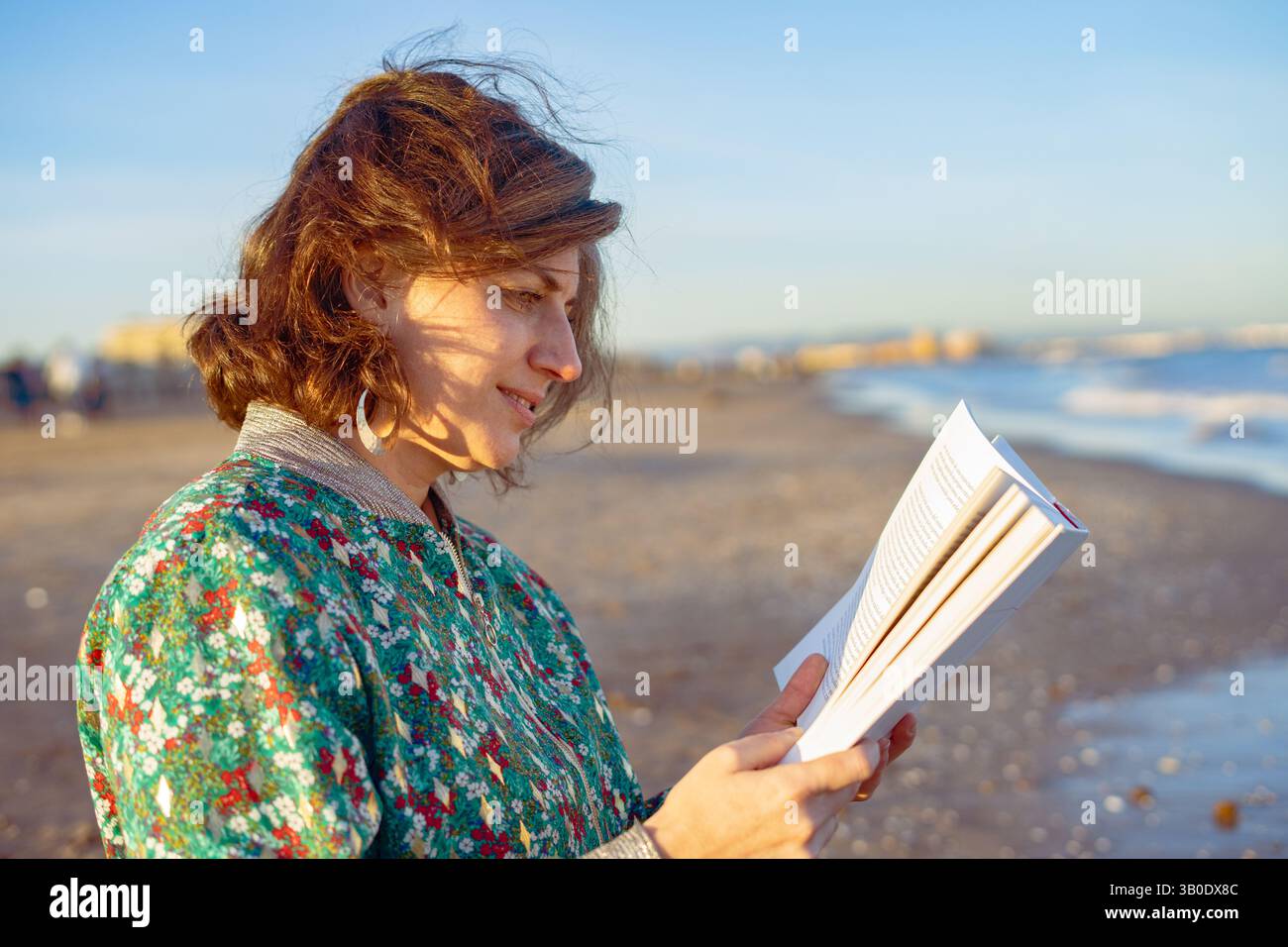Giovane donna in piedi sulla spiaggia al tramonto, immersa in un libro, assaporando la tranquillità e la bellezza del momento mentre le onde si infrangono dolcemente sulla riva Foto Stock