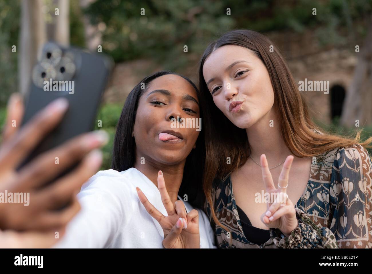 Due giovani donne che creano volti divertenti e segnali di pace mentre scattano un selfie con uno smartphone in un parco vivace, irradiando gioia e amicizia spensierata Foto Stock