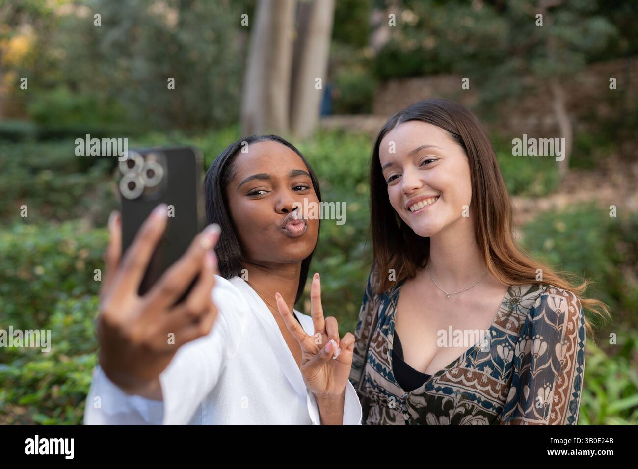 Due amiche stanno facendo volti divertenti e gesti delle mani mentre fanno un selfie con uno smartphone in un parco pubblico Foto Stock