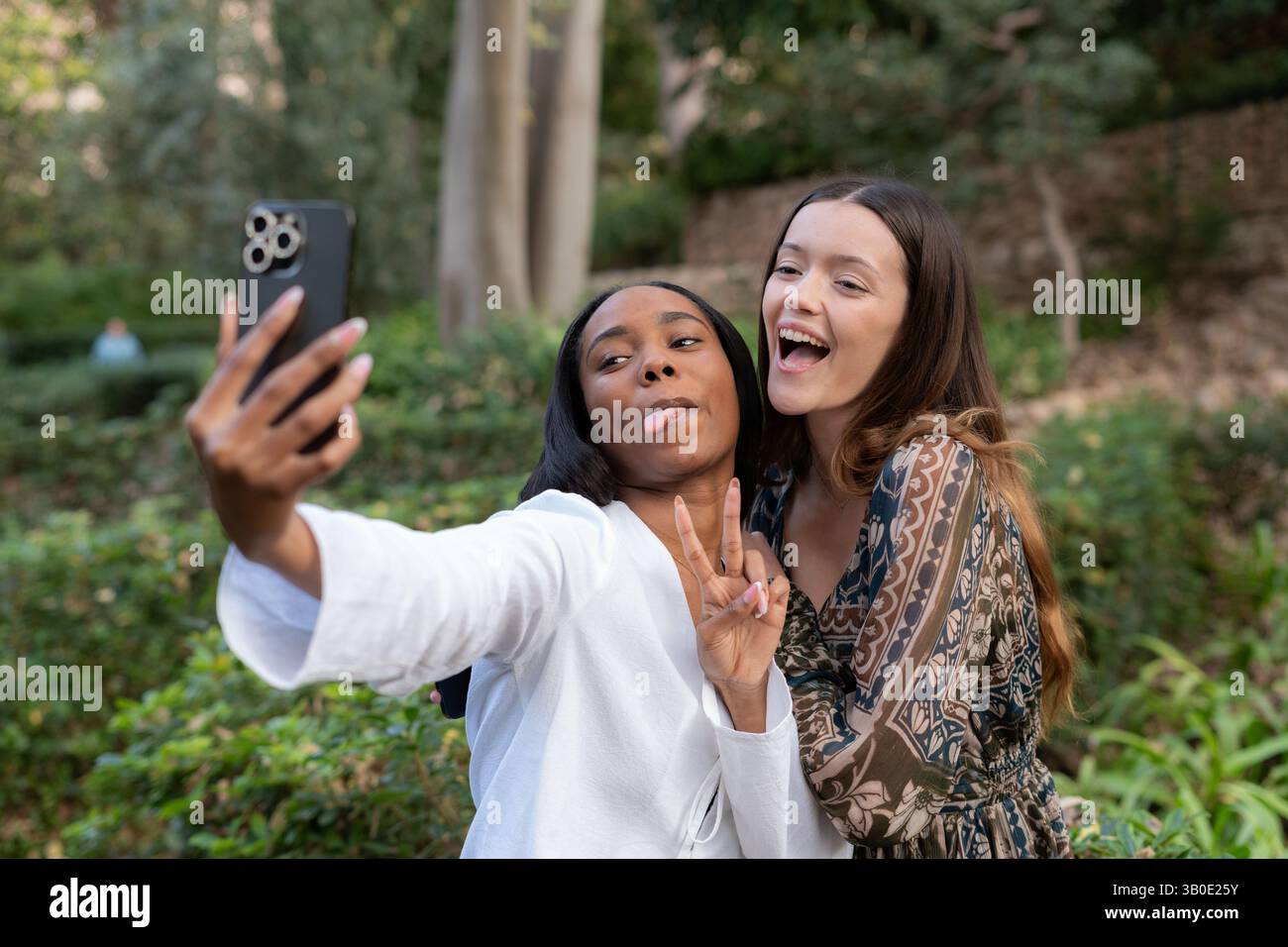 Due amiche multi-etniche si stanno facendo un selfie in un parco, si tirano fuori la lingua e fanno il segno della vittoria con le dita Foto Stock