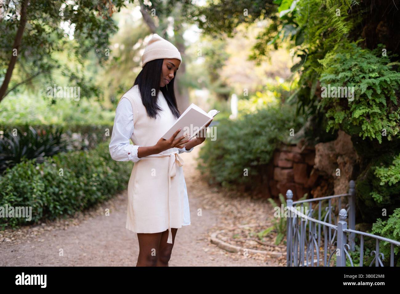 Elegante giovane donna nera che si diverte a leggere un libro mentre si trova su un sentiero in un tranquillo giardino botanico, circondato da una vegetazione lussureggiante, immerso in Foto Stock