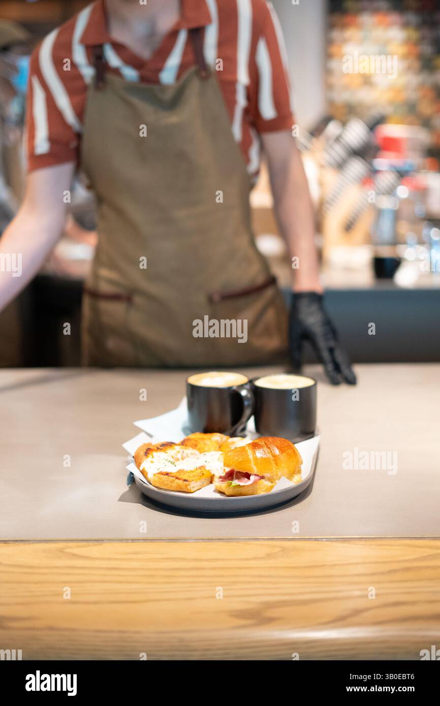Il barista serve panini gourmet freschi per la colazione con caffè artigianale in un ambiente alla moda, catturando l'atmosfera accogliente del brunch e la presentazione del cibo Foto Stock