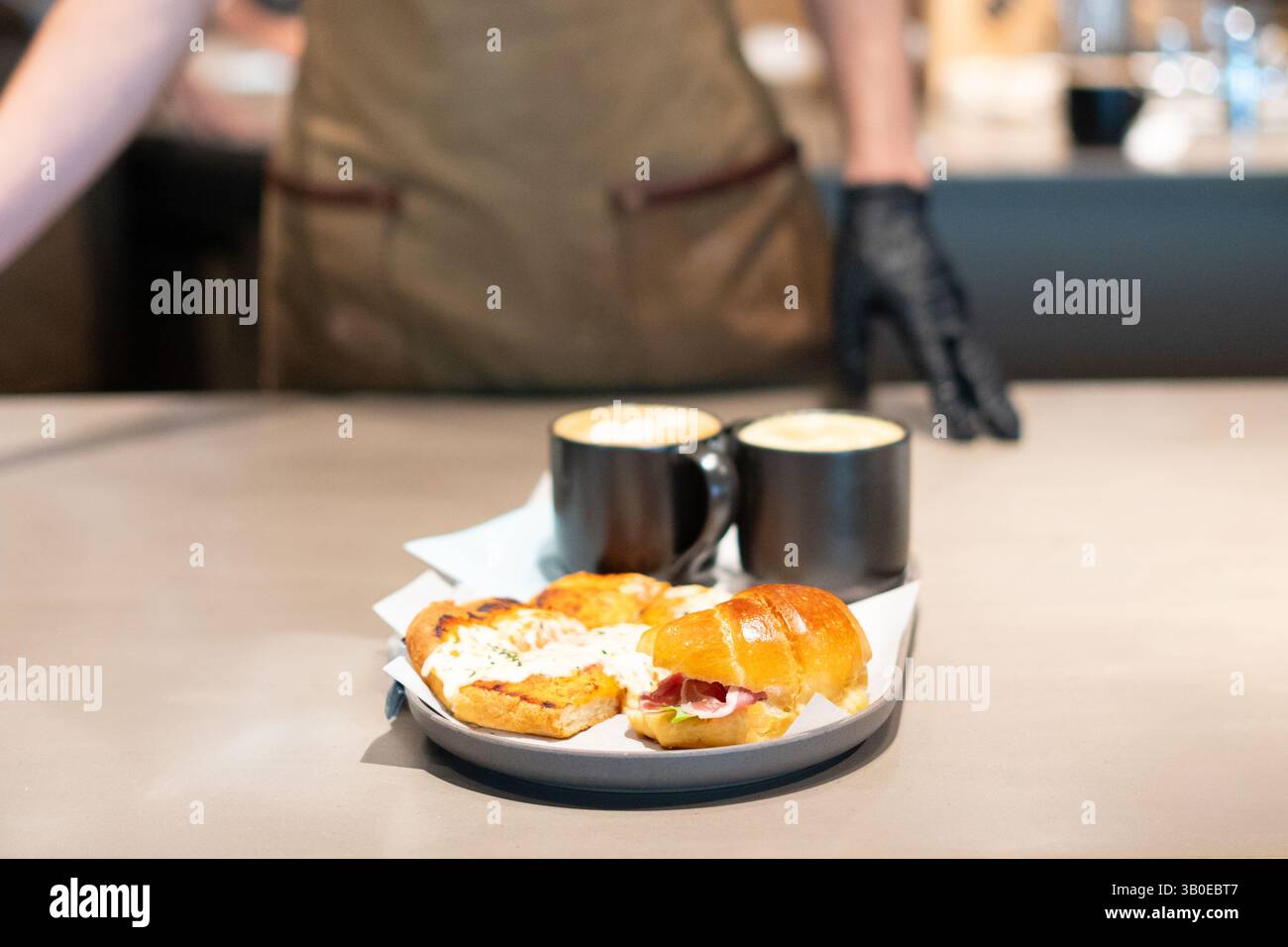 Il barista serve panini gourmet freschi per la colazione con caffè artigianale in un ambiente alla moda, catturando l'atmosfera accogliente del brunch e la presentazione del cibo Foto Stock