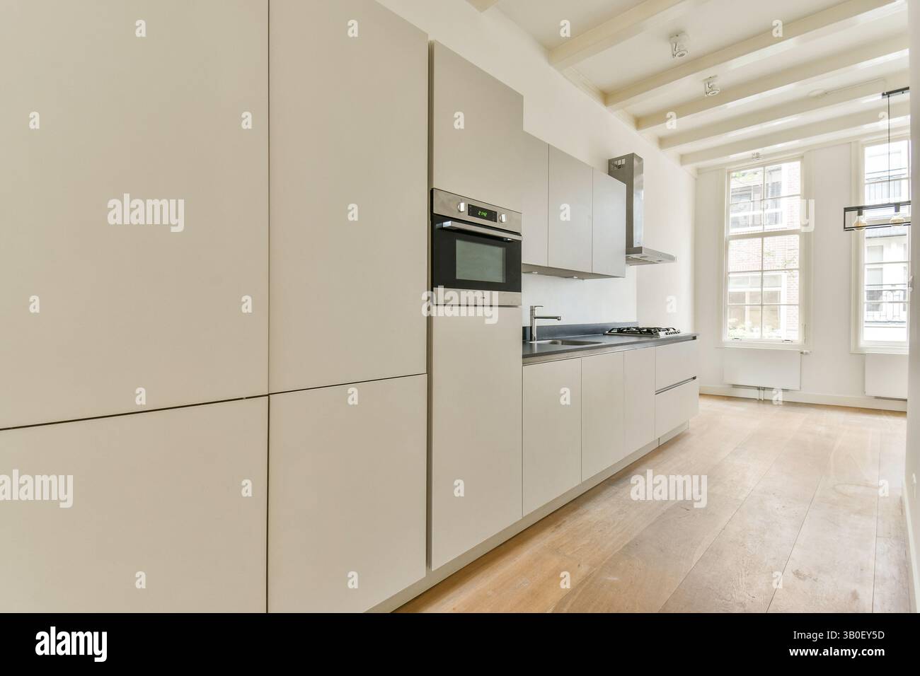 Una cucina elegante e moderna dal design minimalista, caratterizzata da mobili pallidi, pavimento in legno e grandi finestre che illuminano lo spazio con luce naturale. Foto Stock Una cucina elegante e moderna dal design minimalista, caratterizzata da mobili pallidi, pavimento in legno e grandi finestre che illuminano lo spazio con luce naturale. Foto Stock