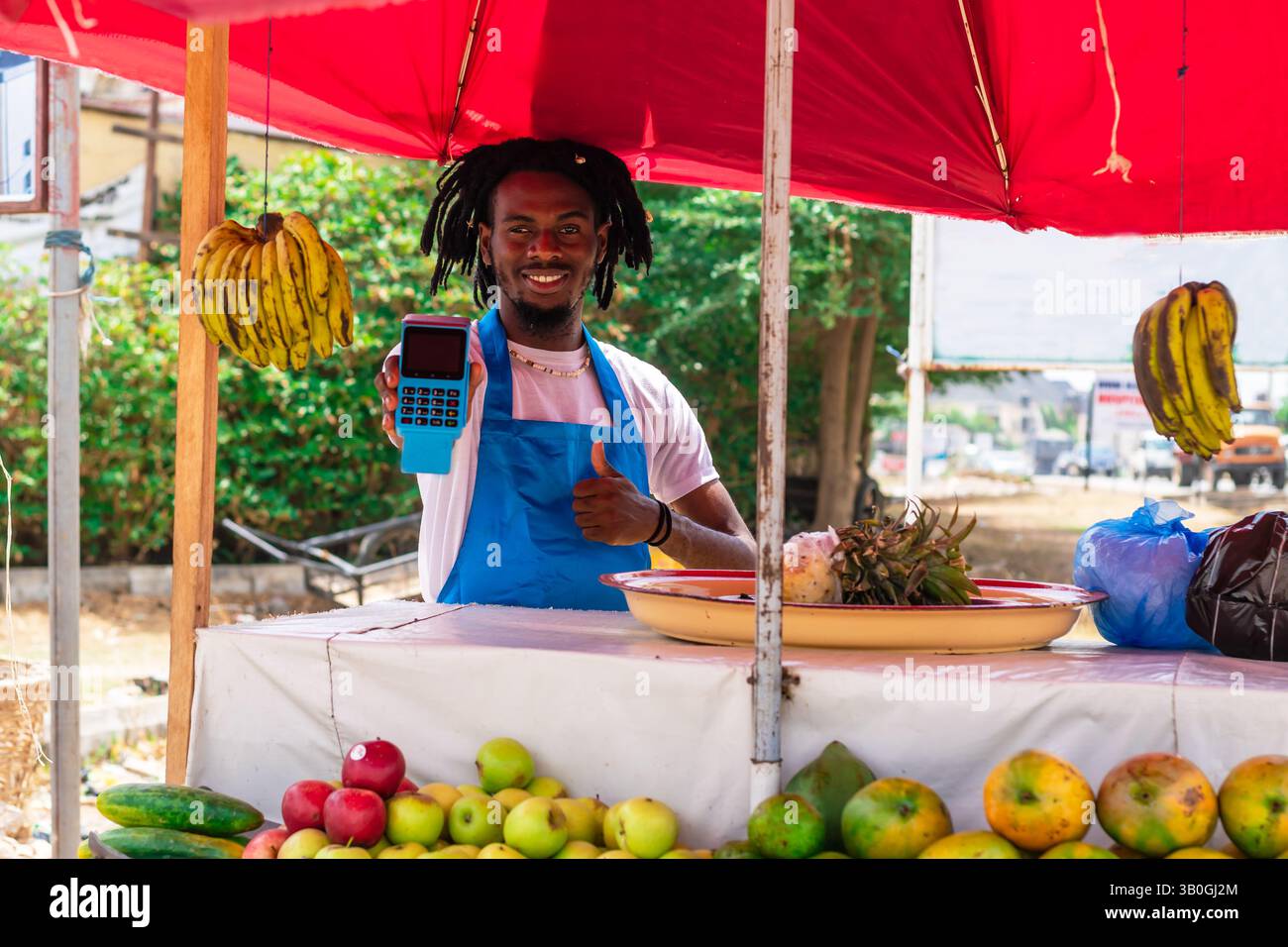 Giovane uomo d'affari africano che indossa grembiule sfocato utilizzando un metodo di pagamento moderno, con un terminale POS e un telefono cellulare Foto Stock