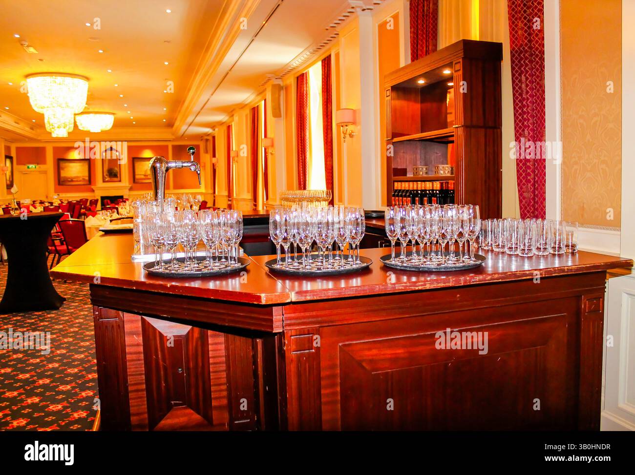 Un'area bar splendidamente arredata con diversi vassoi di bicchieri scintillanti, allestita in un'elegante sala banchetti. La calda illuminazione e il lussuoso decor creano Foto Stock