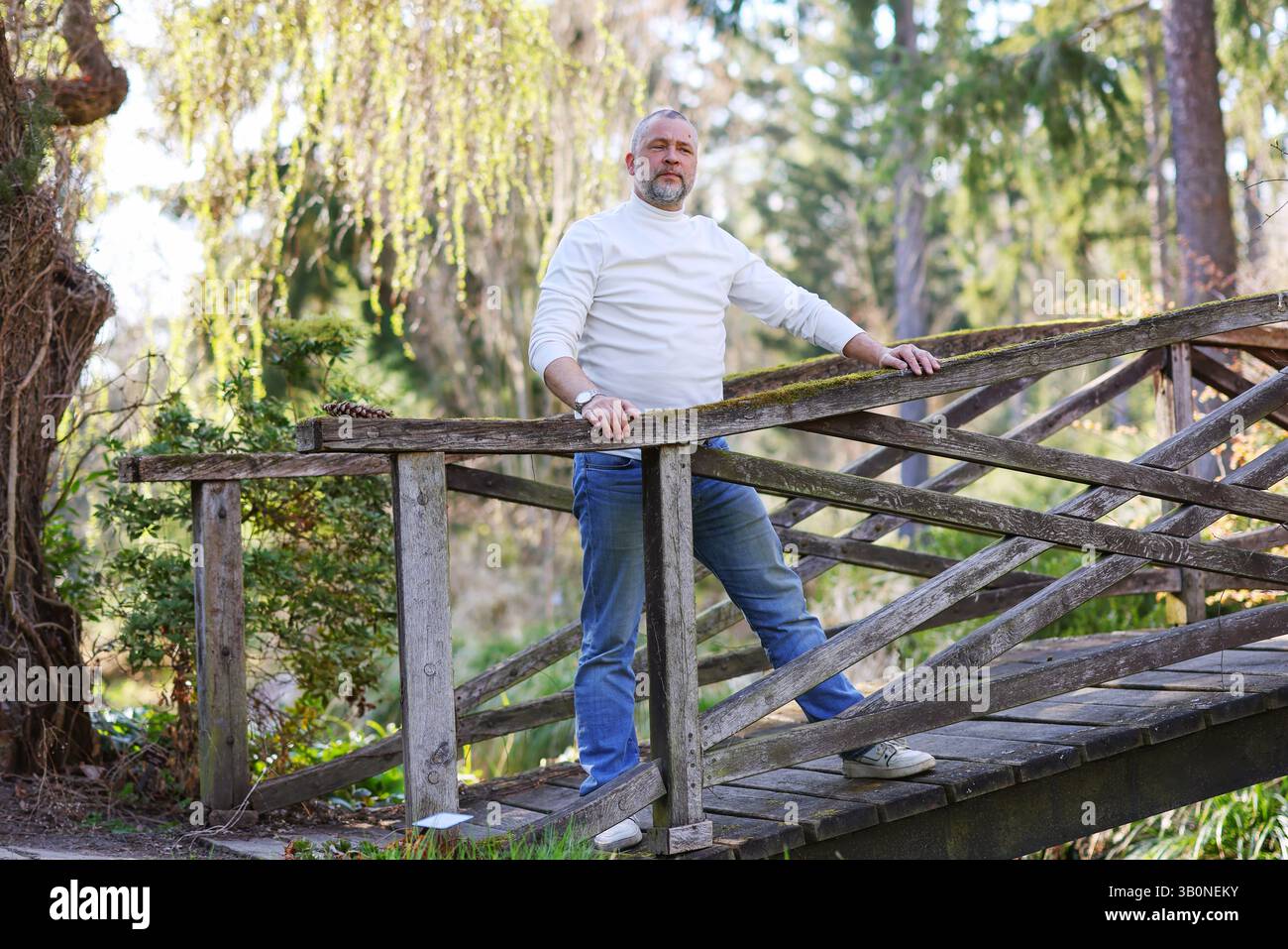 L'uomo barbuto in un abbigliamento casual posa all'aperto su un ponte intempestivo circondato da alberi e luce del sole. Foto Stock