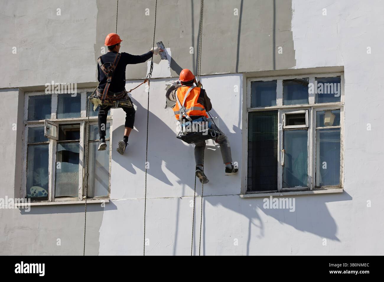 I lavoratori riparano il muro dell'edificio mentre sono appesi a funi di sicurezza. I costruttori dipingono la facciata della casa, i lavori di costruzione e riparazione Foto Stock
