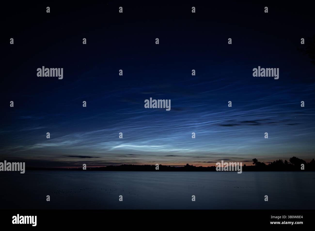 Delicate nuvole nocciolenti si estendono attraverso un cielo crepuscolo sopra un tranquillo lago estivo. Le loro scintillanti sfumature bluastre creano un contrasto etereo Foto Stock