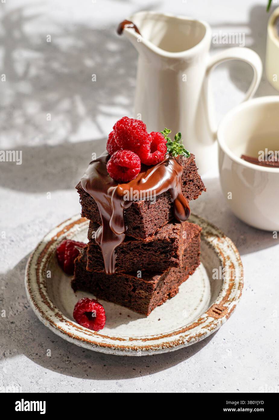 brownie al cioccolato ricoperti di cioccolato fuso lucido e lamponi freschi, serviti su un piatto di ceramica rustica. Il dessert è posto su un li testurizzato Foto Stock