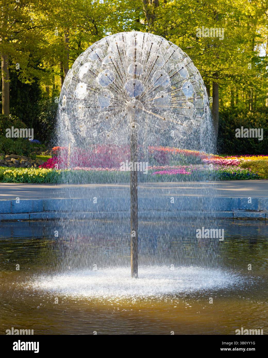 Moderna fontana sferica che spruzza gocce in uno stagno dei Giardini Keukenhof, Paesi Bassi, con aiuole colorate e alberi sullo sfondo. Foto Stock