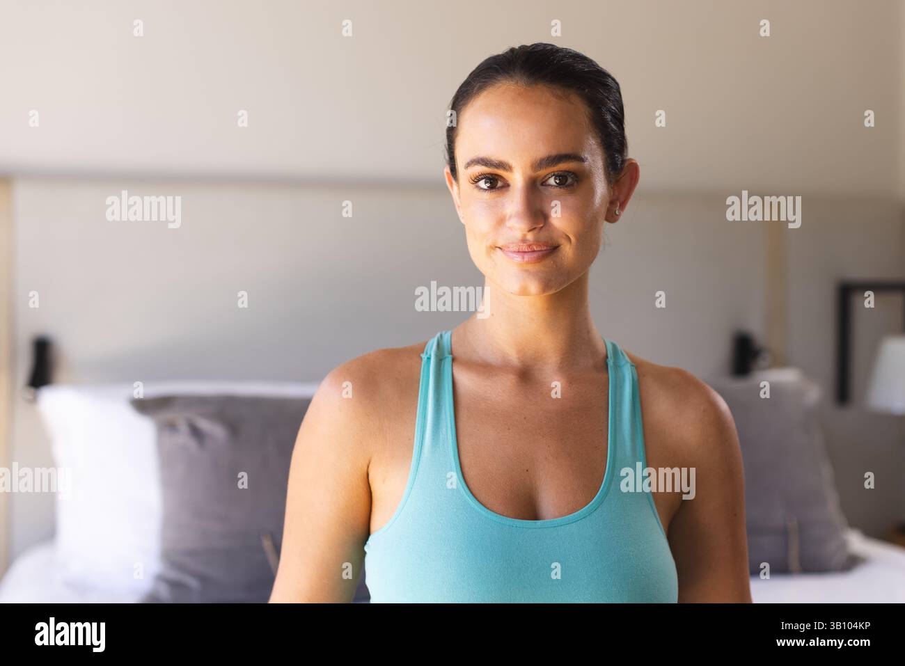 Donna in piedi sorridente delicatamente mentre indossa una canotta turchese di fronte al letto, con lampade da comodino Foto Stock