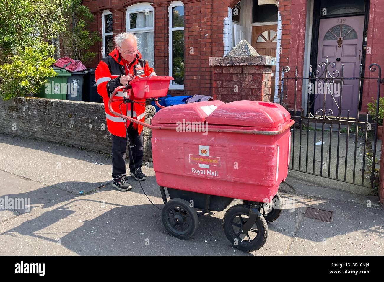 Un postino della Royal mail e il suo carrello a ruote per i pacchi in consegna in un'area residenziale di Cardiff. 24 aprile 2025. Foto Stock